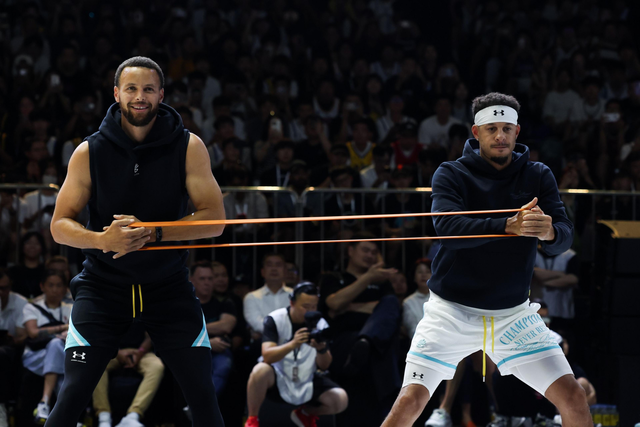 Two athletes stretch an orange resistance band between them while standing in a sports arena filled with spectators.